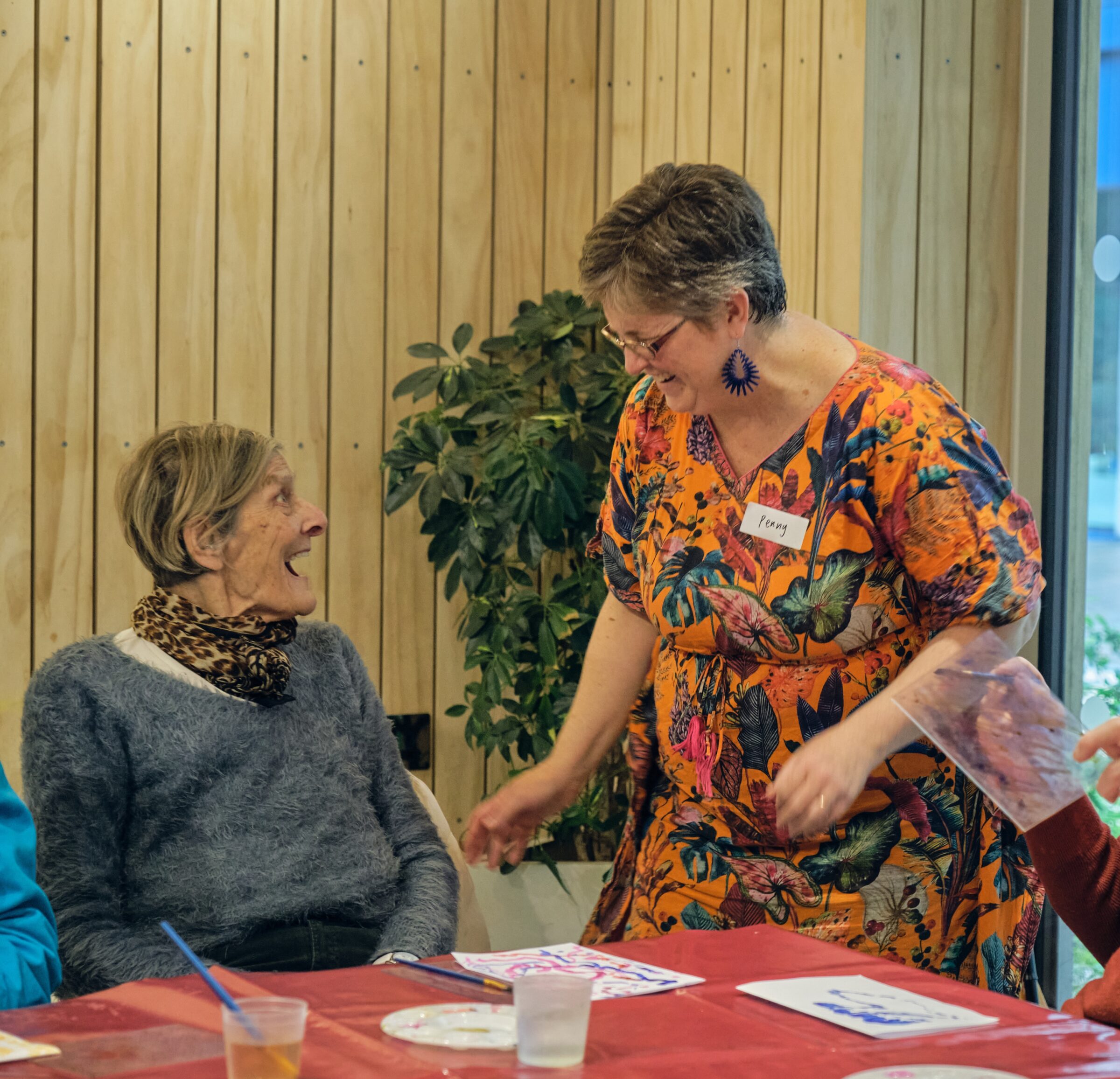 An older woman is sitting and laughing up at a woman in an orange floral dress