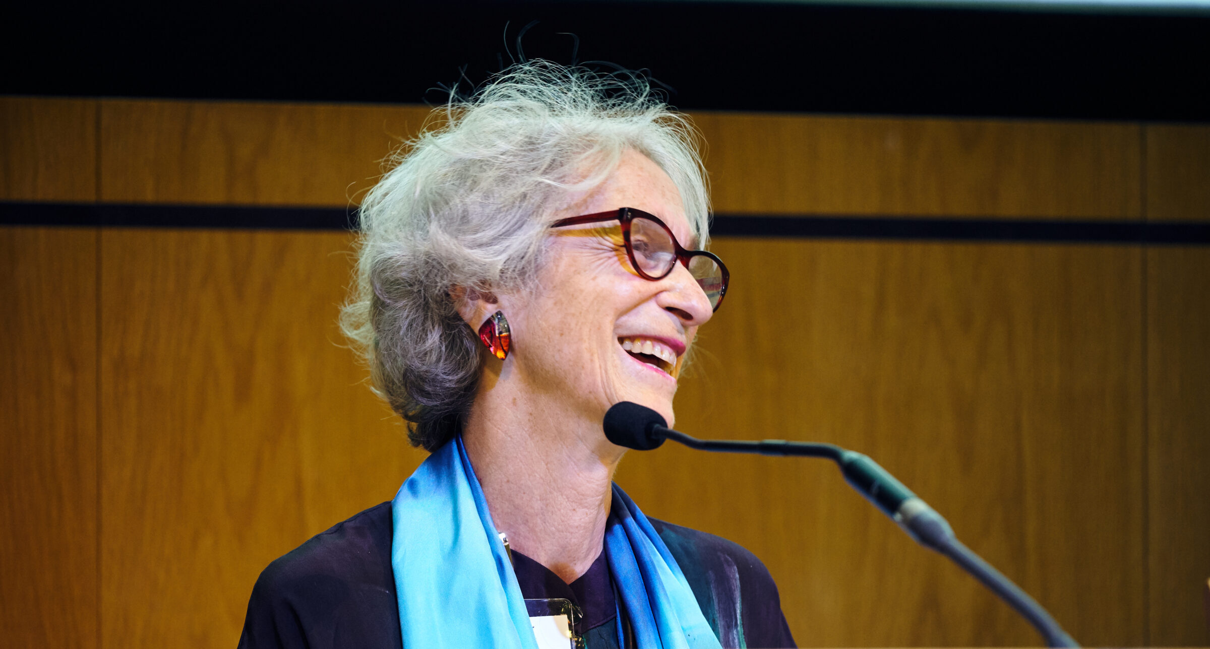 Veronica is behind a podium and microphone, smiling towards the audience