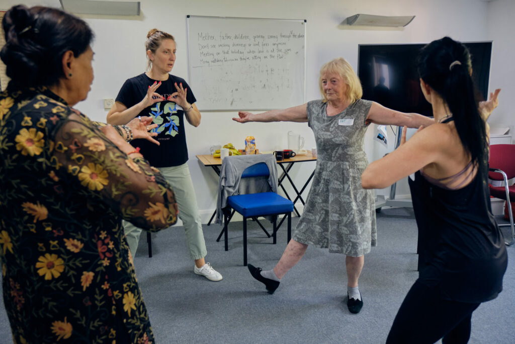 Four women are standing in a circle. One woman is striking a dance pose with her arms out, the others are trying different hand movements.