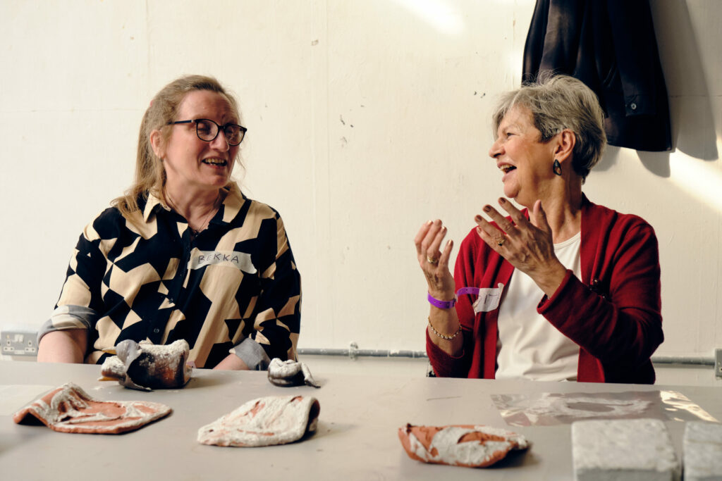 Two women are sitting next to each other, folded clay in front of them, as they chat and smile.