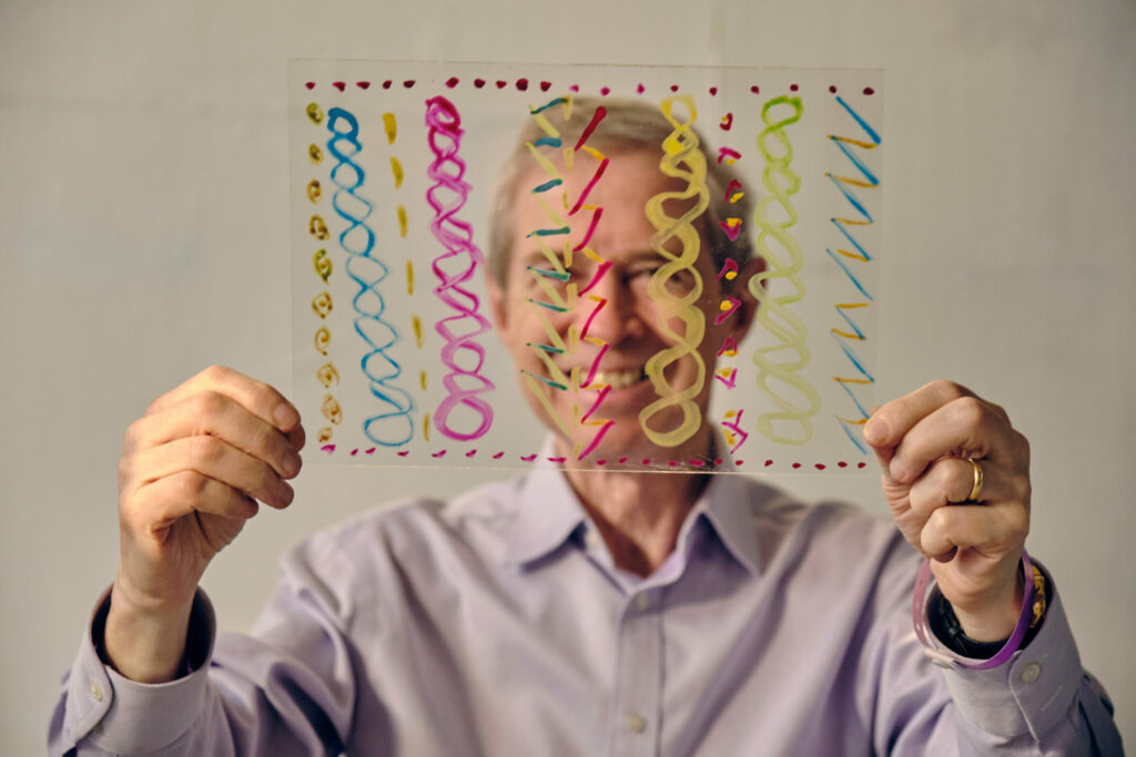 A man is holding up a clear piece of plastic that is covered in colourful lines and patterns, you can see his broad smile through the plastic.