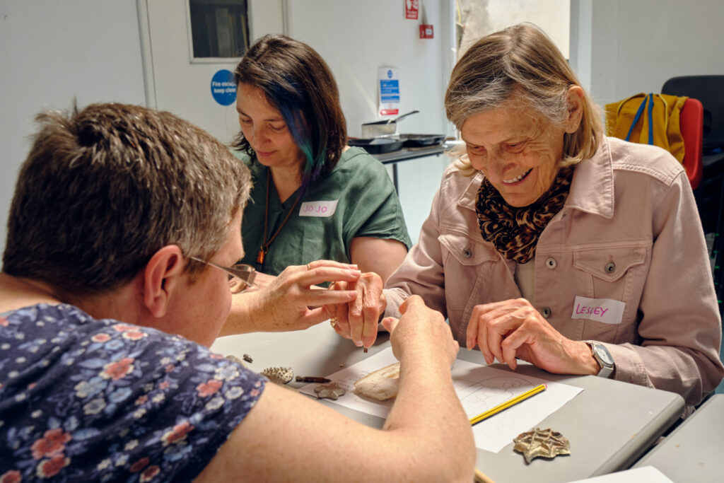 Three women sitting around a table. One woman is holding the hands of an older woman as they work to carve into cuttlefish. The older woman is smiling.