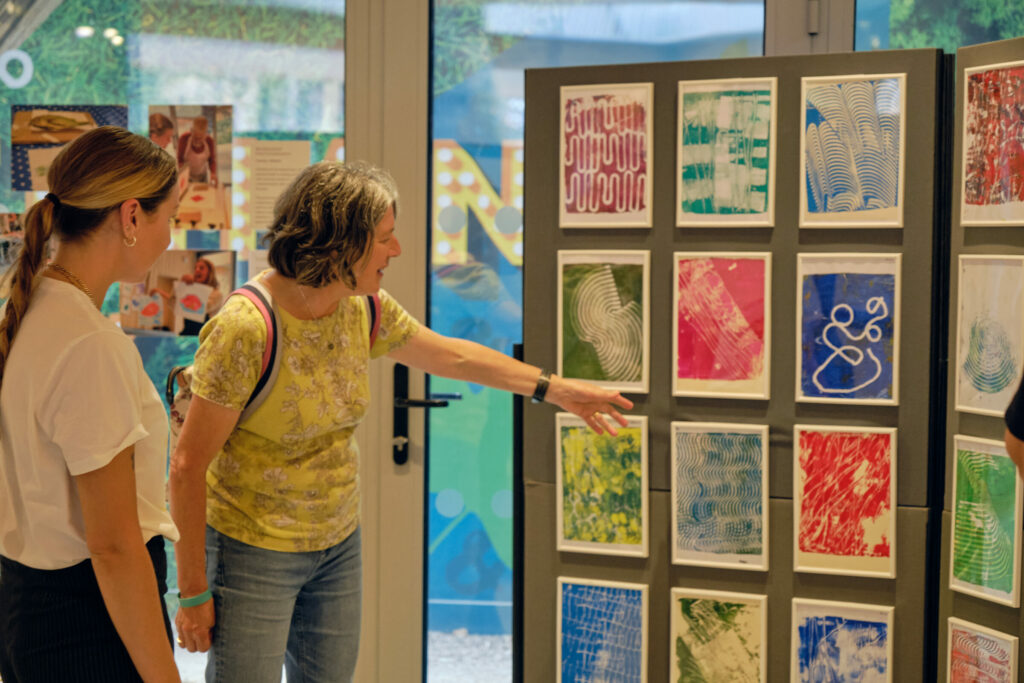 Two women are looking at colourful, framed gel-plate prints