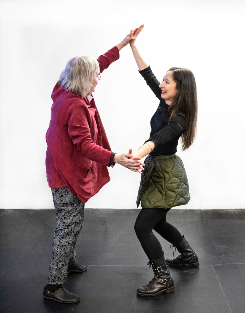 A younger woman and an older woman are smiling as they dance together with their hands pressed together.