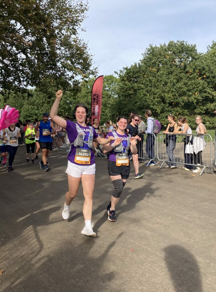 Two young women are running together in purple t-shirts, and smiling and waving to people at the side of the road.
