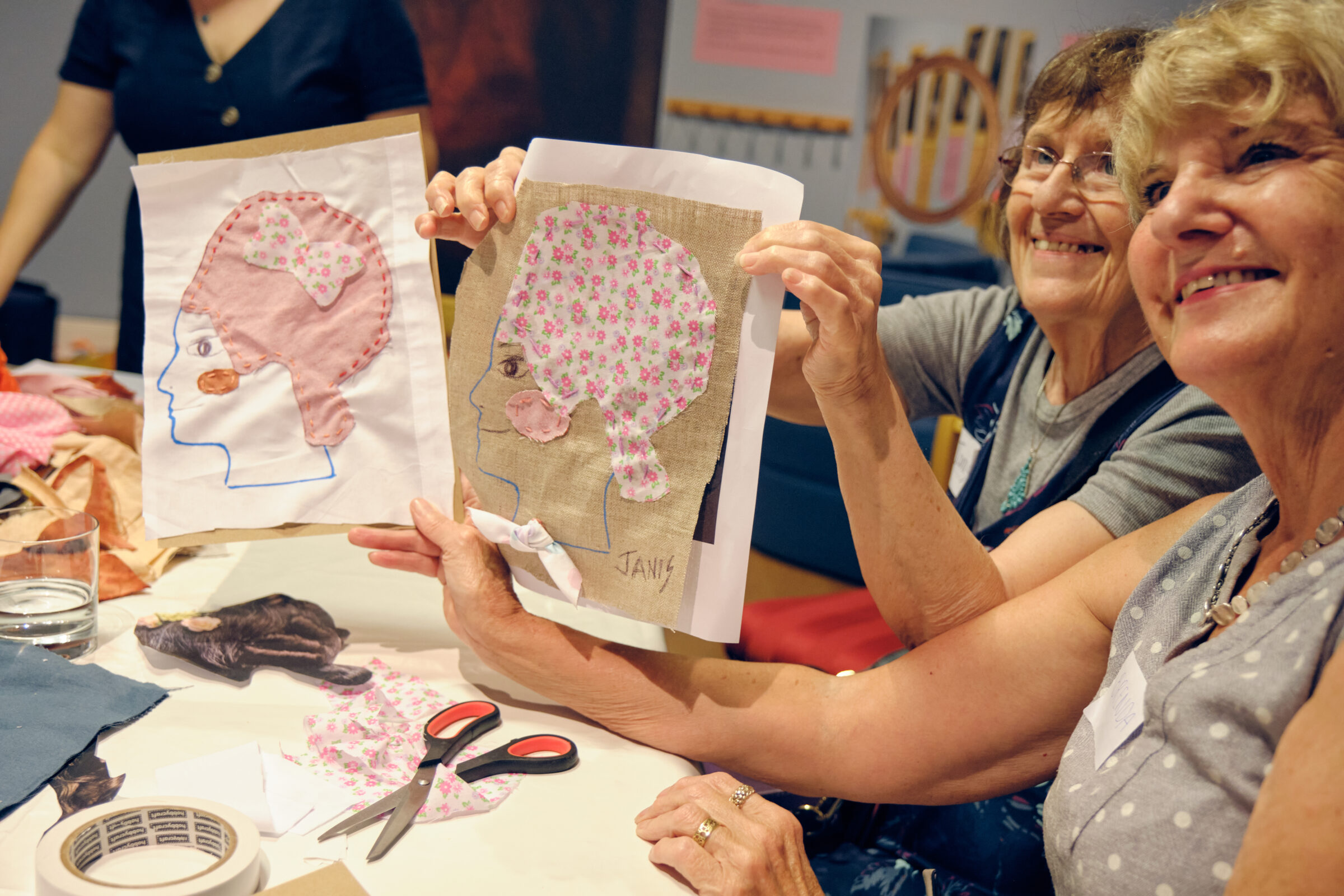 Two older women are sitting next to each other, smiling broadly and holding up pictures of their faces that they have made by sewing different fabric together.