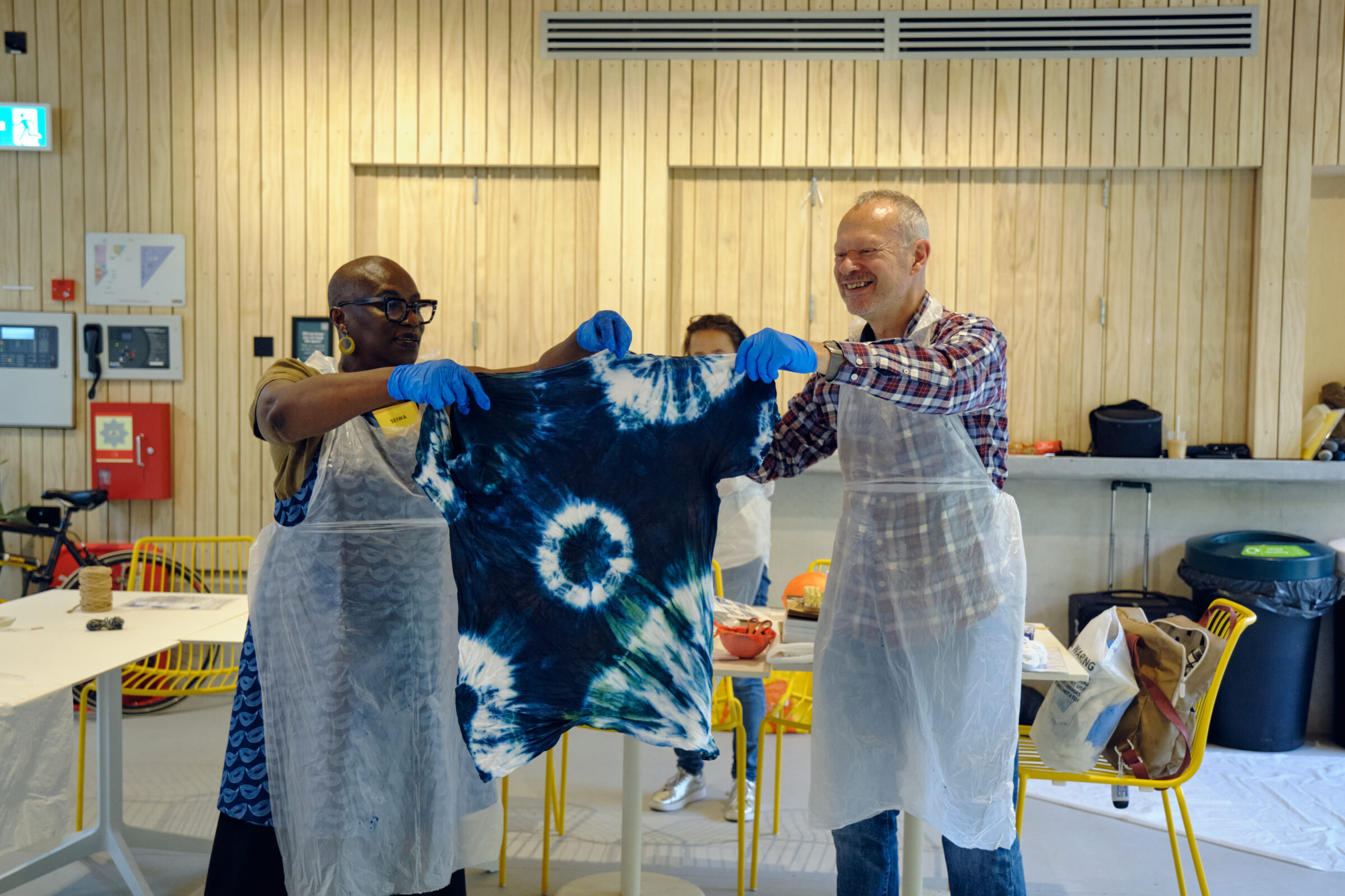 A man and a woman in white aprons are holding between them a t-shirt tie-dyed with indigo and white circular patterns. They are both smiling.