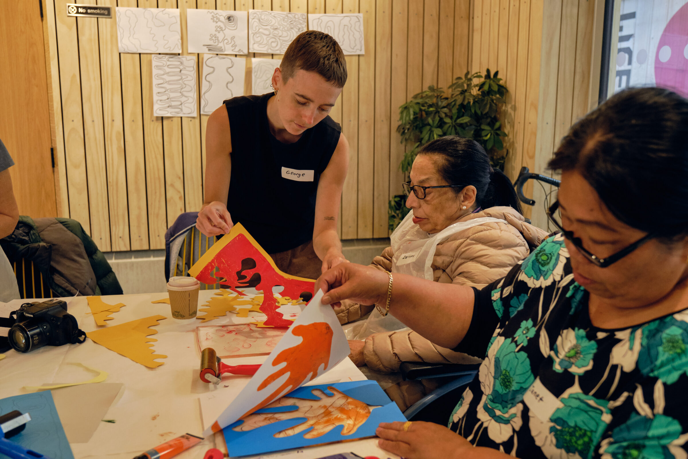 A young woman is standing and helping an older woman with cutting colourful paper into detailed shapes. Another woman has printed a shape and is seeing her new art.