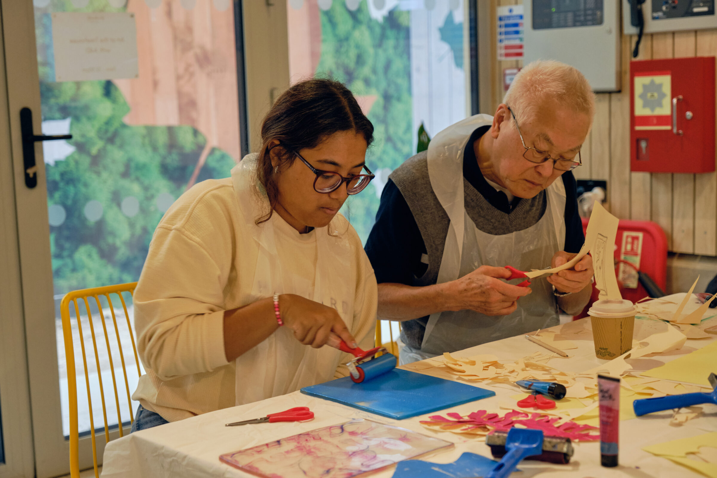 Two people are concentrating on creating. A younger woman is rolling blue ink onto a picture, and an older man is cutting out fine details into paper.
