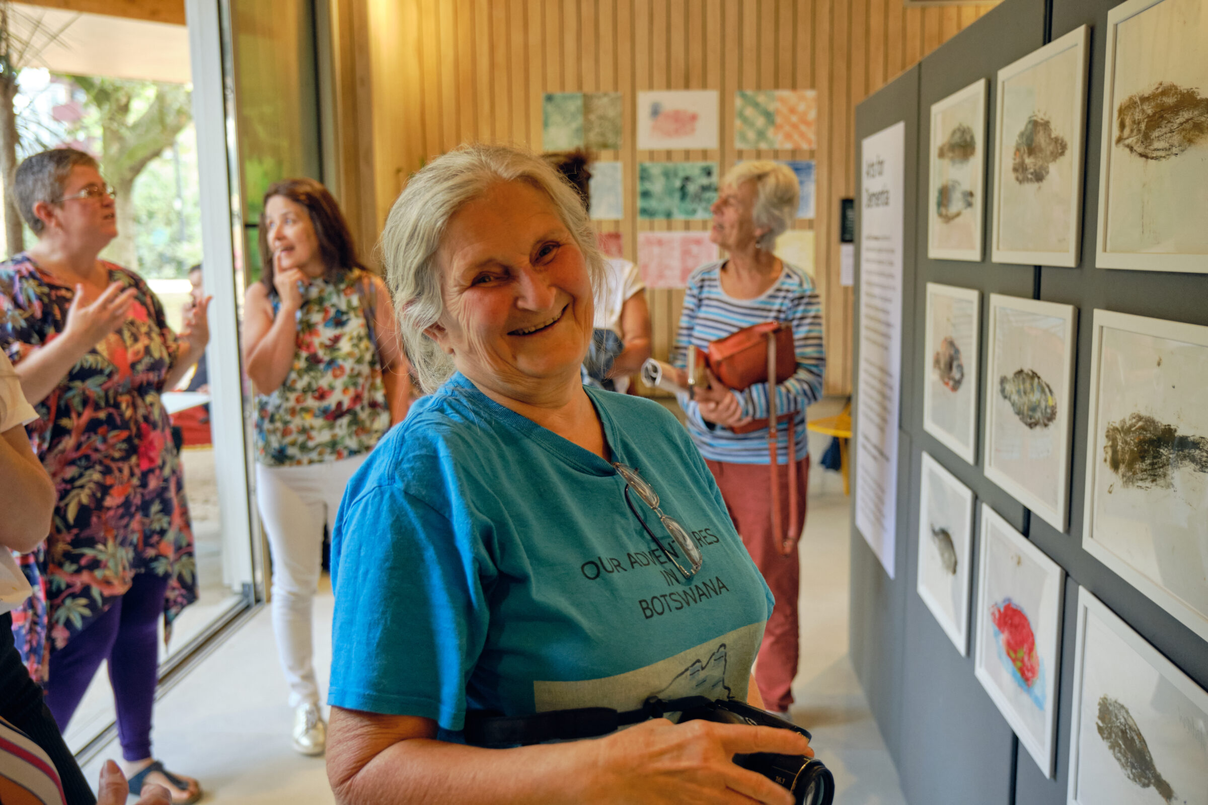 Gill is smiling broadly at the camera, standing beside prints of fish created by her, Dave and other participants in our Treehouse print-making programme. Other people are talking in the background.