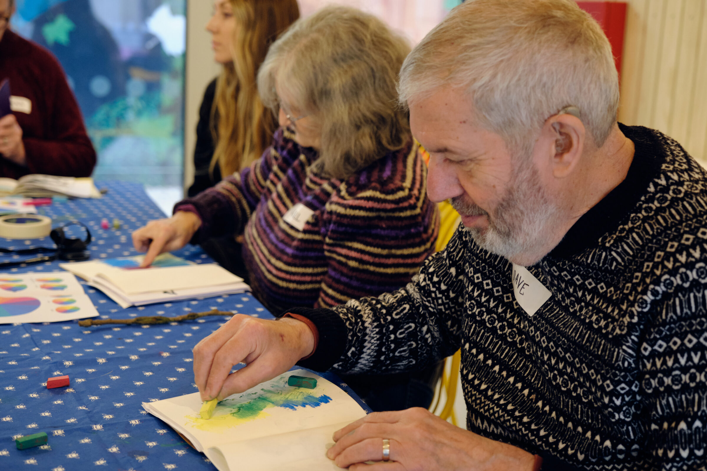 Dave is holding a yellow pastel and adding to a picture that already has blue and green and pencil lines. He is smiling gently. In the background Gill is using her finger to blend pastel colours.