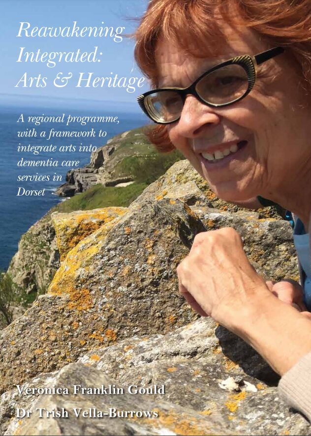 The words 'Reawakening Integrated: Arts & Heritage. A regional programme with a framework to integrate arts into dementia care services in Dorset' are on an image of a woman in glasses looking out to sea on a rocky coastline.