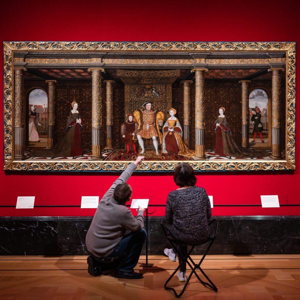Two people looking and one pointing at a large painting depicting a royal scene from Tutor times by Holbein in a red walled gallery.