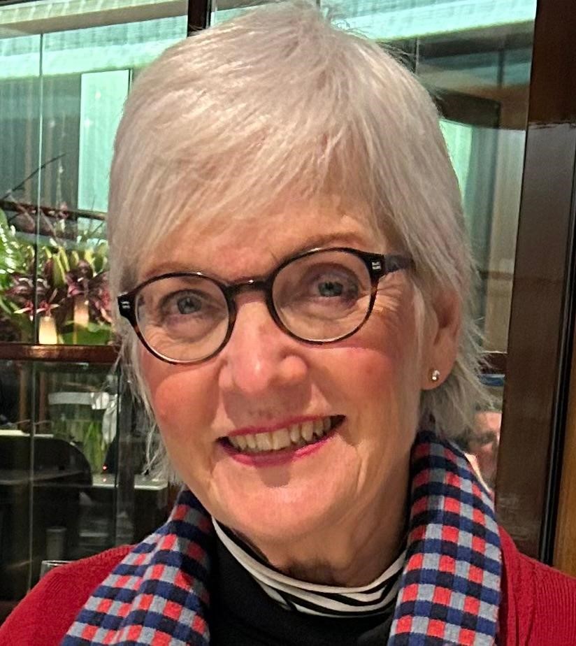 Lori is an older woman with short, white hair, black-framed glasses, and wearing a red jacket with a red, blue and black checked collar. She is smiling at the camera.