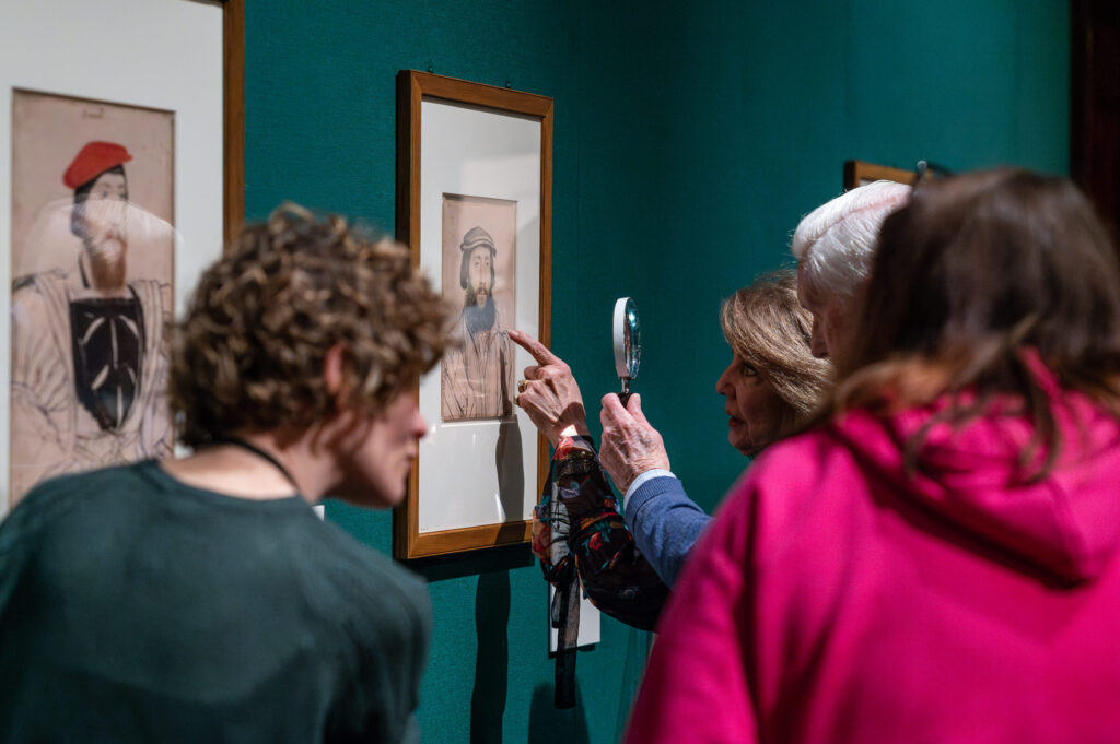 4 people are looking at a portrait by Holbein on the wall of a gallery. One person has a magnifying glass to look more closely.