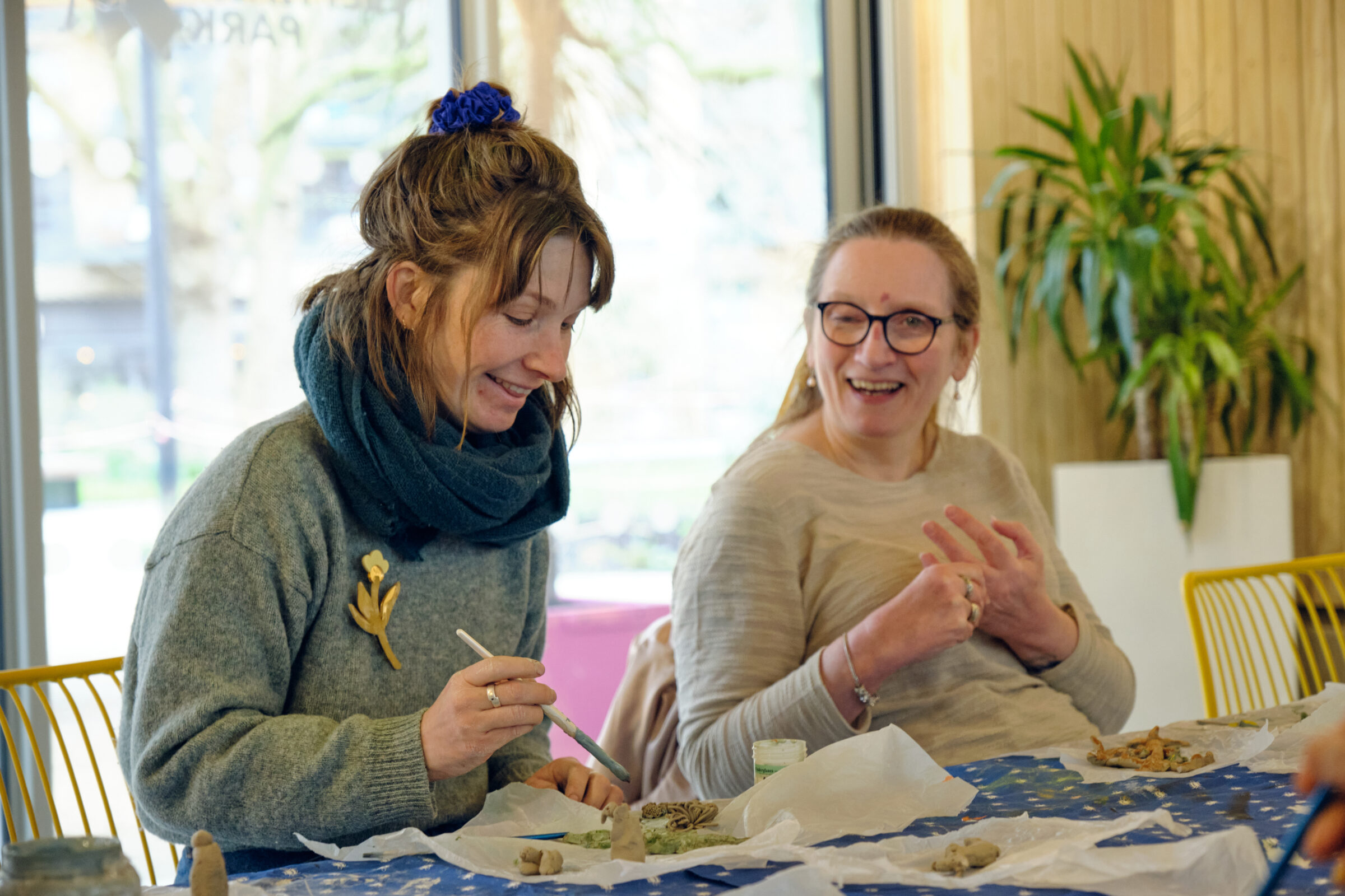 Two women are sitting next to each other, smiling, while painting onto clay items