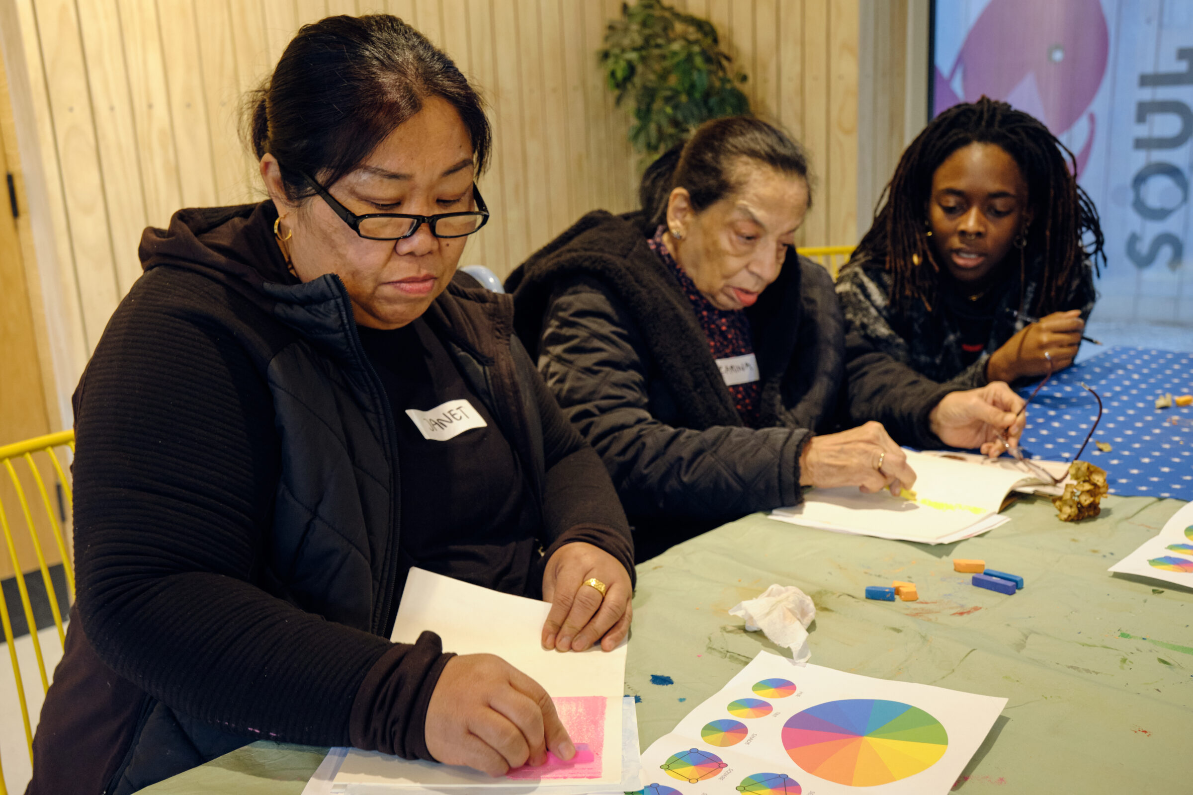 Three women are sitting at a table, using colourful pastels to create shapes.