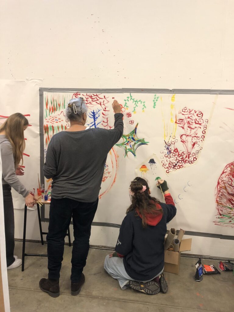 Two people are painting on a huge piece of paper taped to a wall, creating a collaborative piece of art with different colours, shapes and styles.