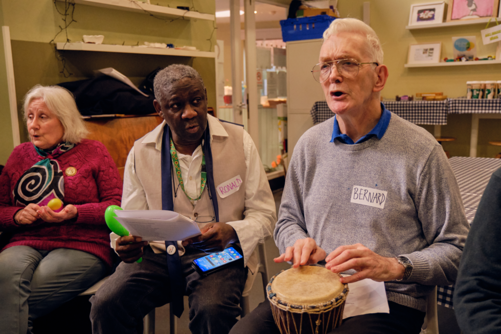 Three people seated next to each other with one playing a drum the other two singing