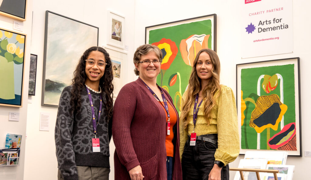 Three women are standing together in front of a brightly coloured painting of large flowers, beside a sign reading 'Arts for Dementia, Charity Partner'