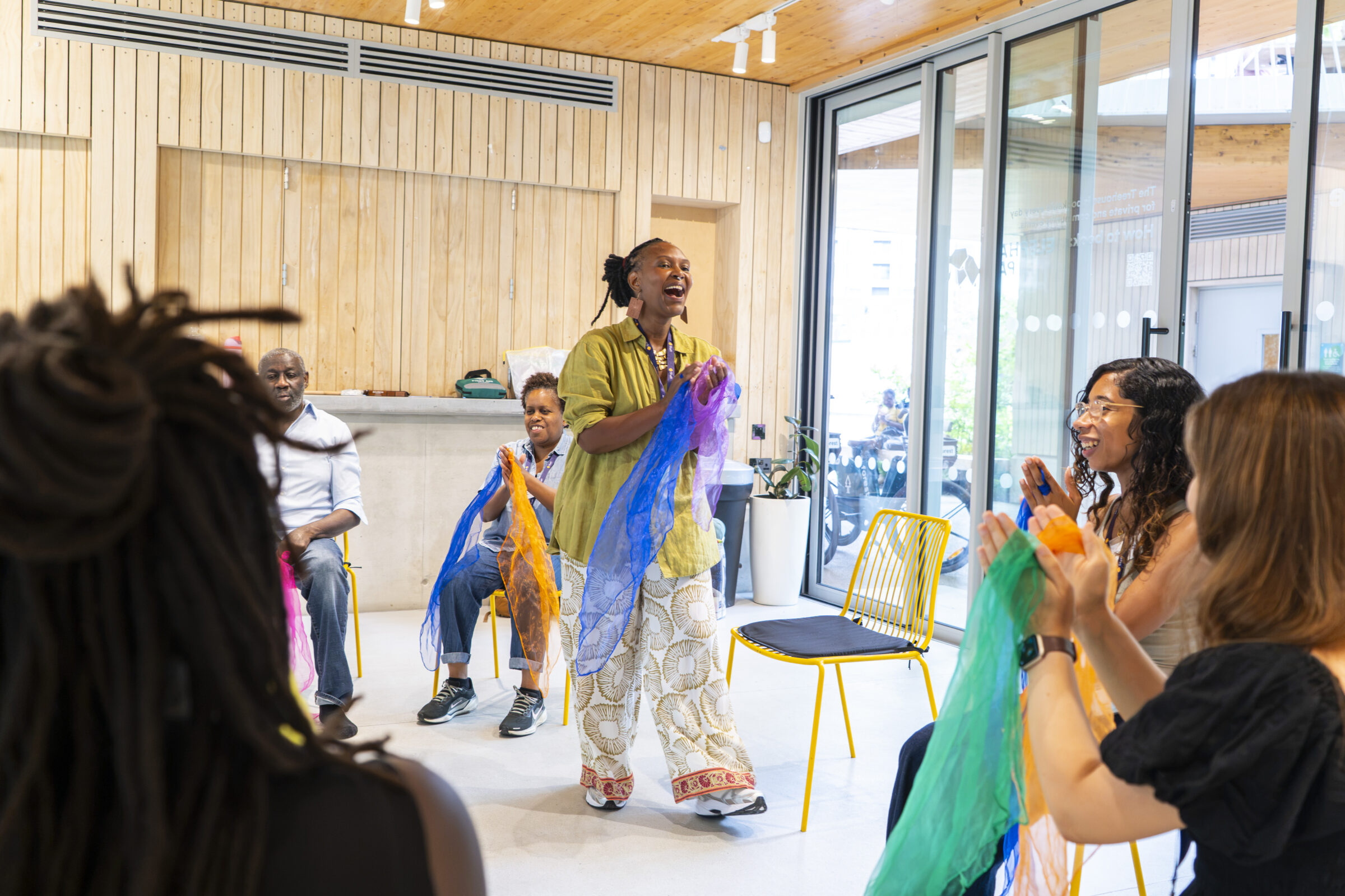 A smiling black woman is standing and smiling, clapping her hands and holding blue and purple floaty scarves. Other people are seated, clapping with scarves of other colours