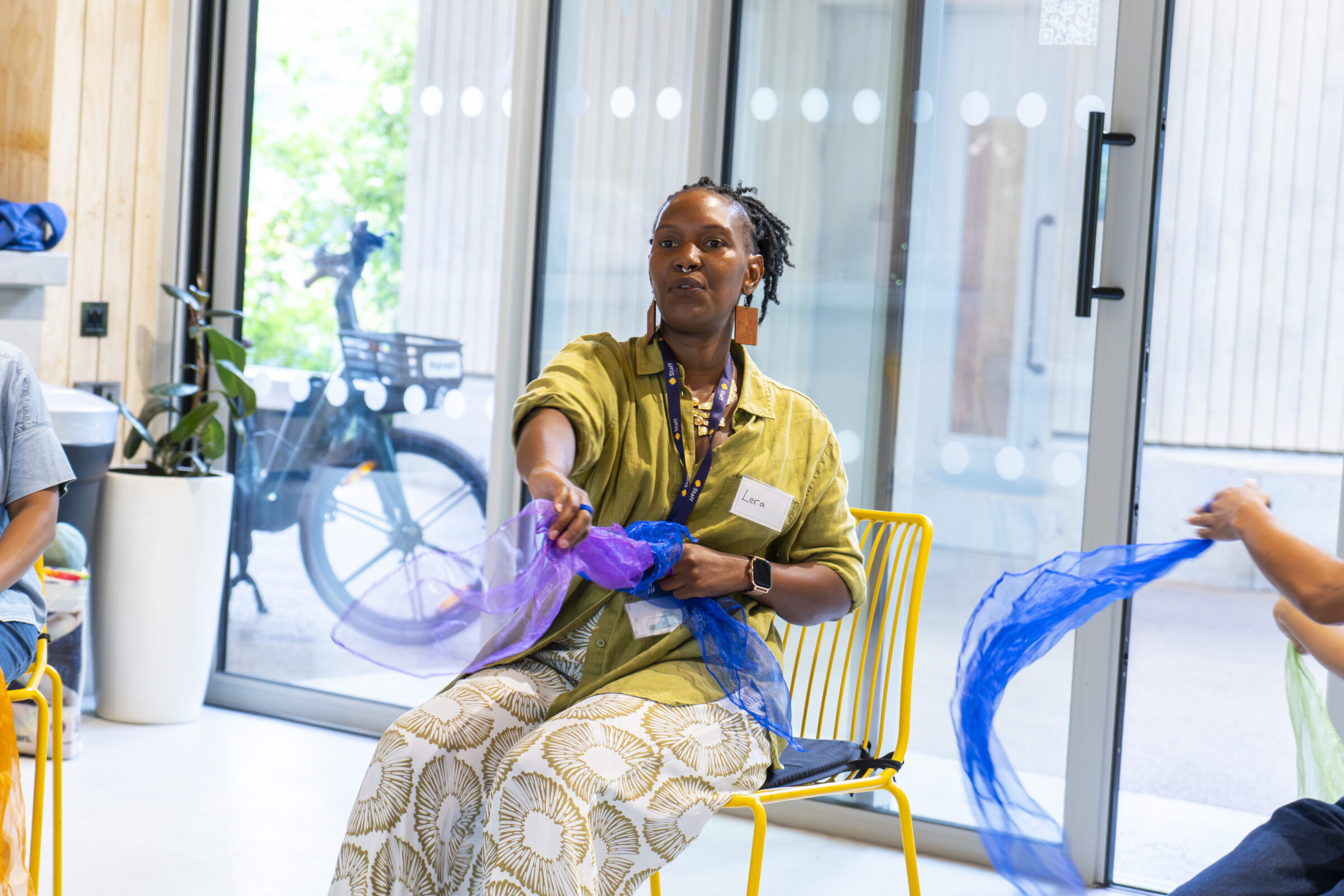 A black woman is sitting and moving with a sheer purple scarf and blue scarf