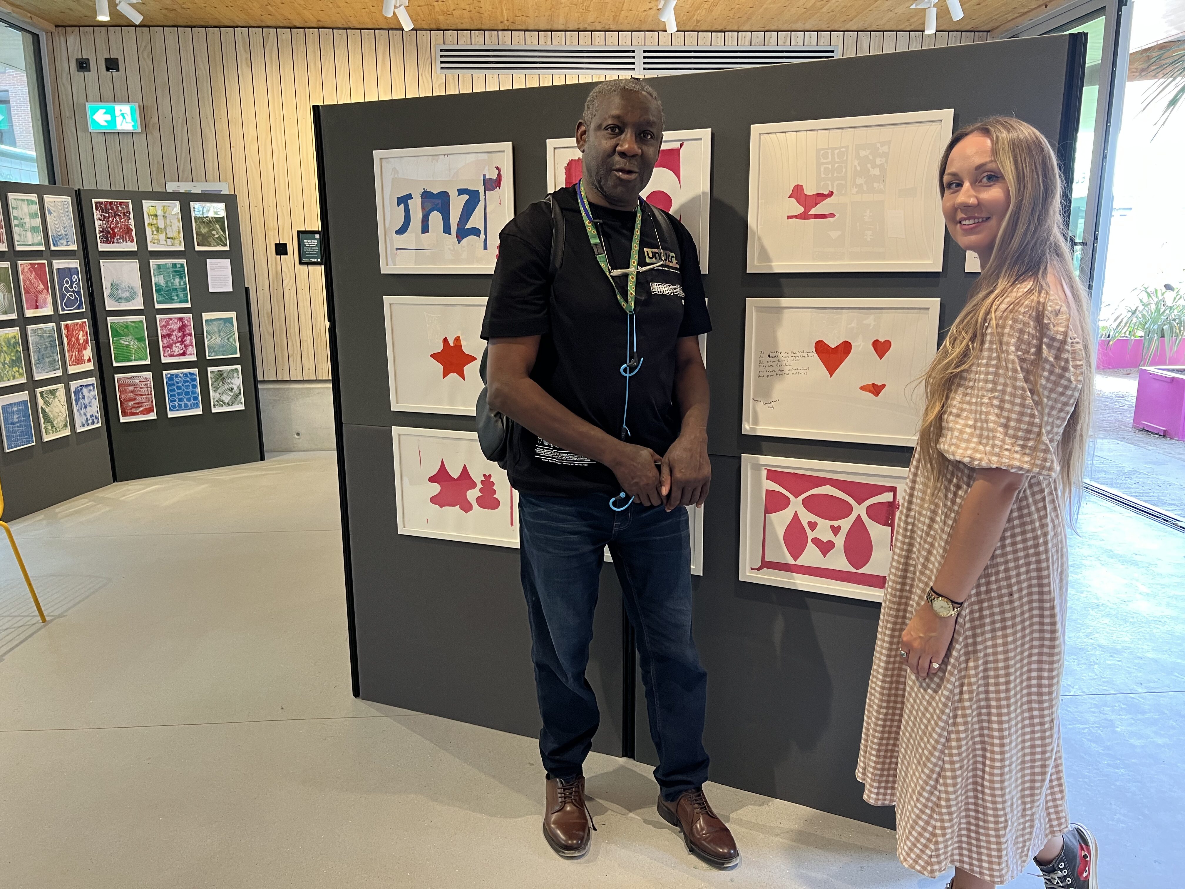 An older black man and a younger white woman stand in front of colourful prints in white frames